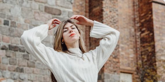 Confident woman in white sweater posing against rustic stone architecture outdoors.