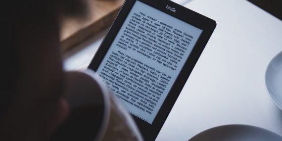 A person reads on a Kindle e-reader while holding a cup of coffee, indoors.